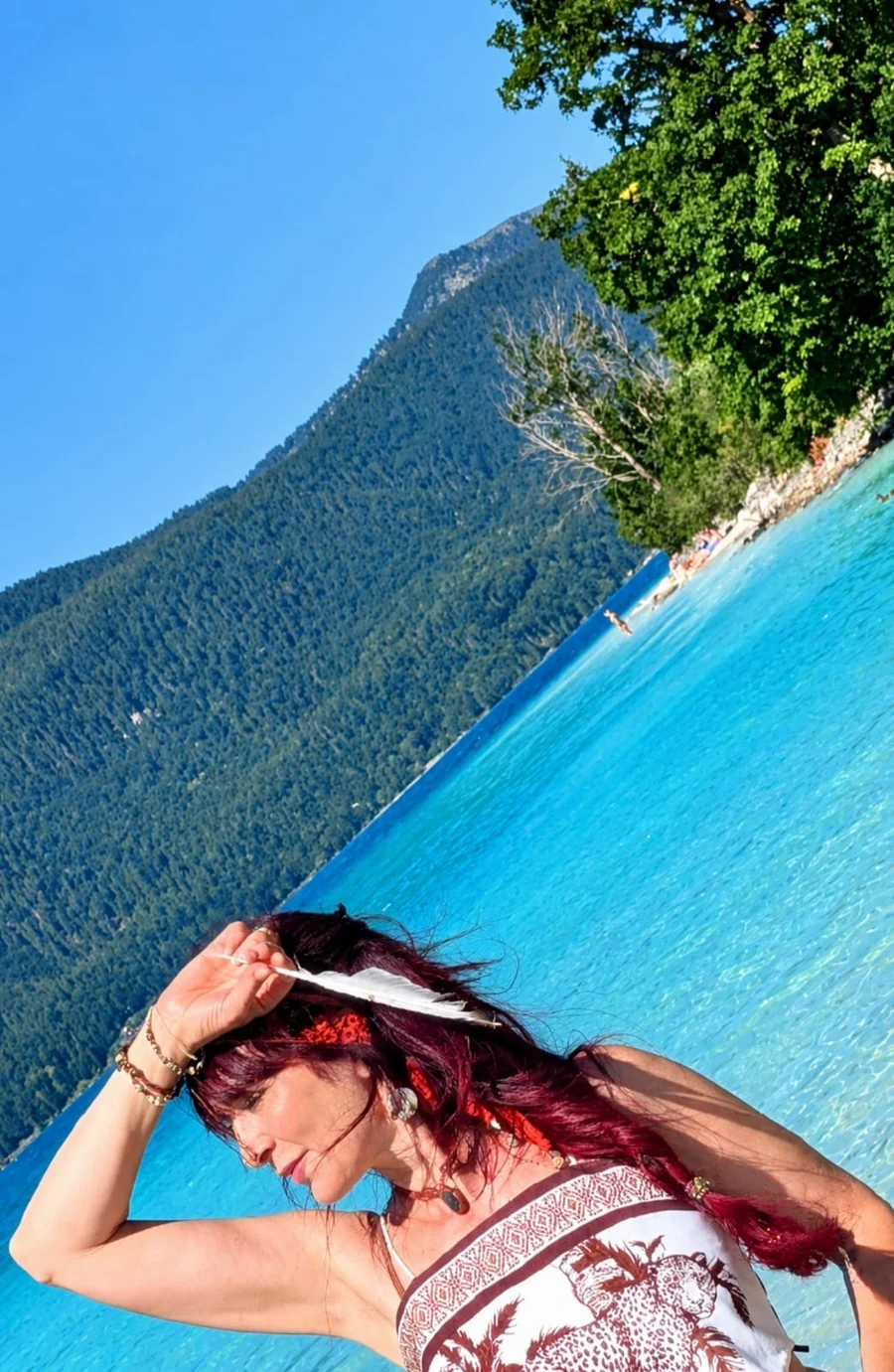 Katharina mit einer weißen Feder im Haar am türkisfarbenen Wasser vor bewaldeten Bergen